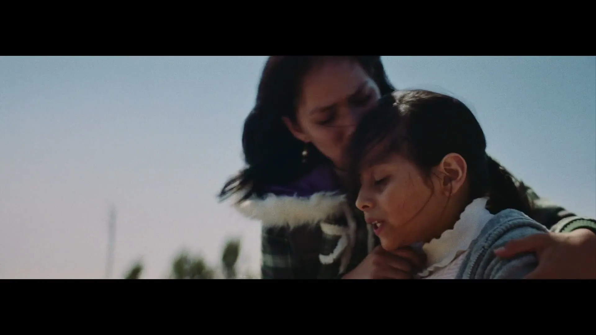A young woman with long dark hair comforts a child, gently touching her face and hair, outdoors under a clear sky.