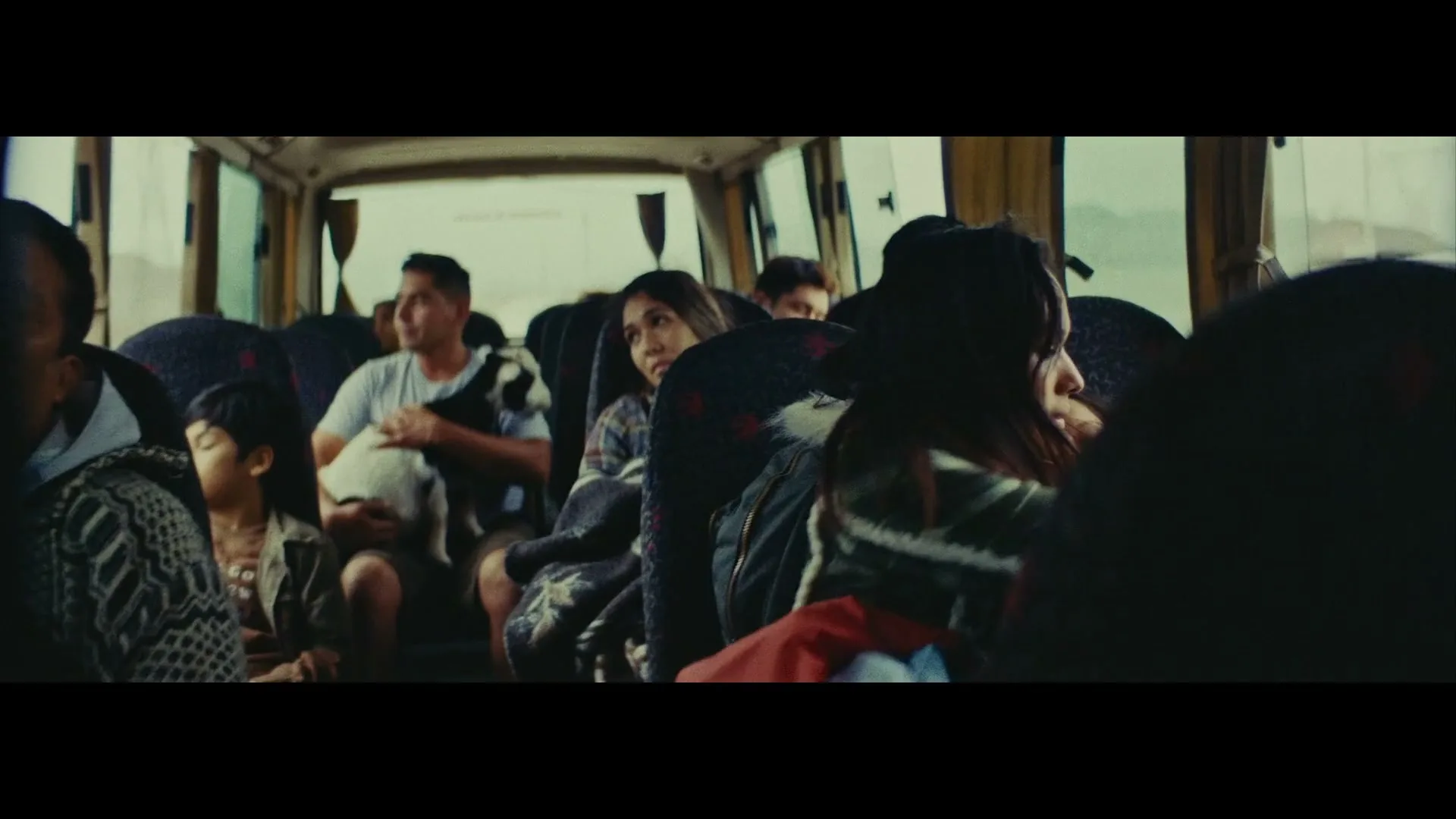 A woman with long dark hair holds a child sleeping on her shoulder, looking out the window of a moving bus with other passengers.