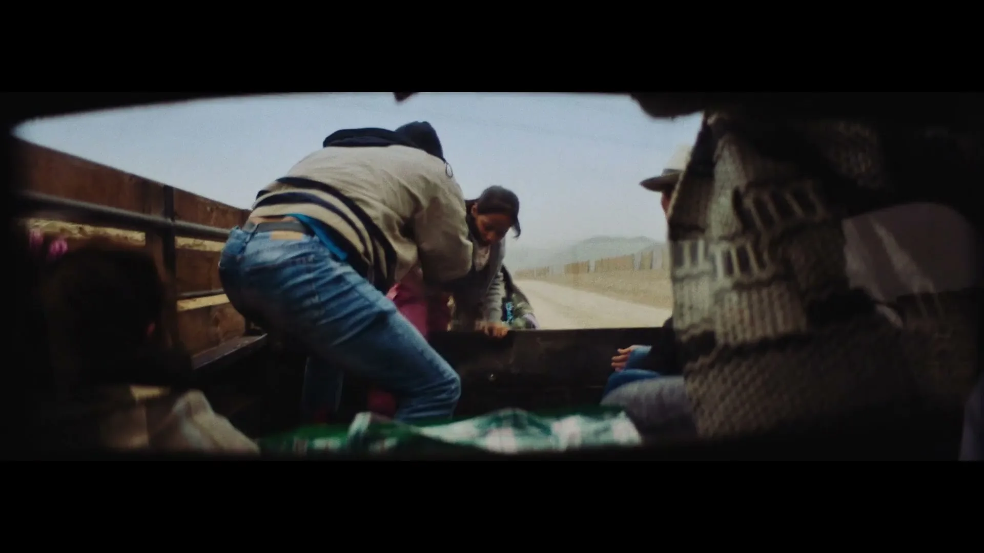 A man in a striped jacket and jeans helps a woman climb into the back of a moving truck on a dusty road.