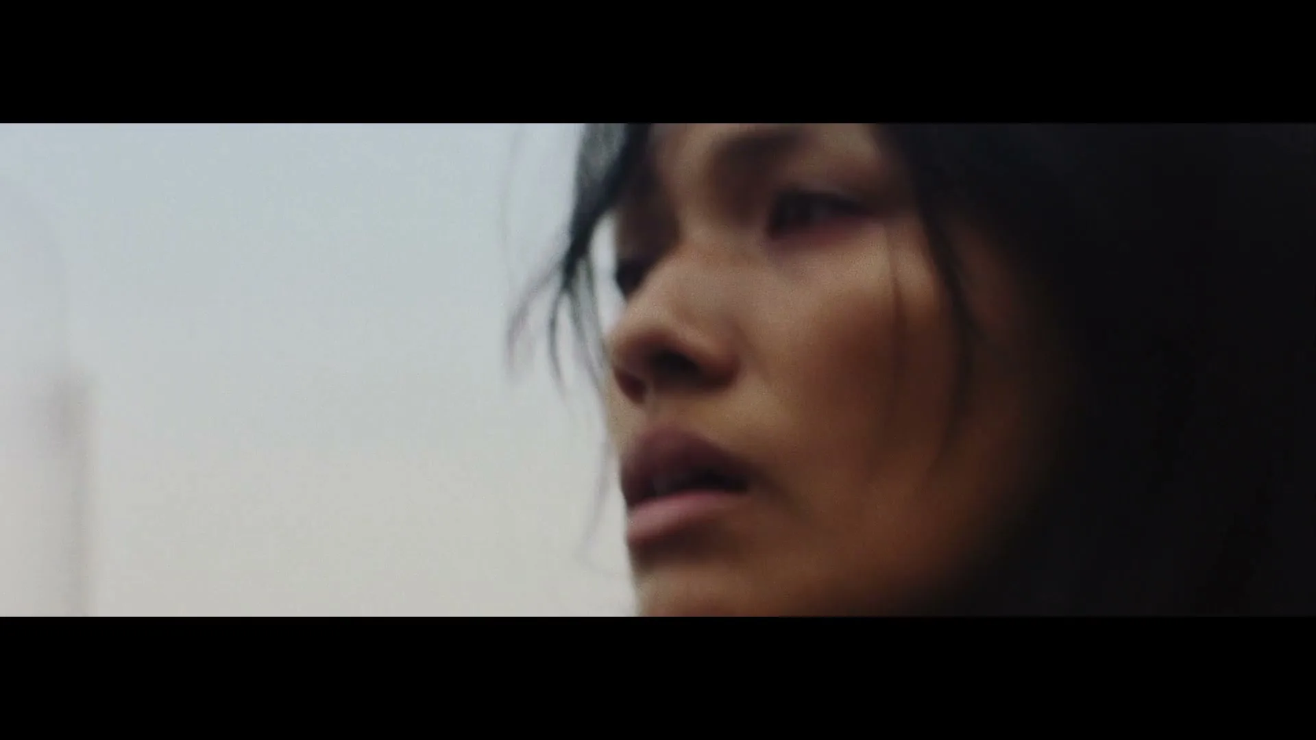 A young woman with dark hair and a somber expression looks off into the distance, with a blurred, light background behind her.