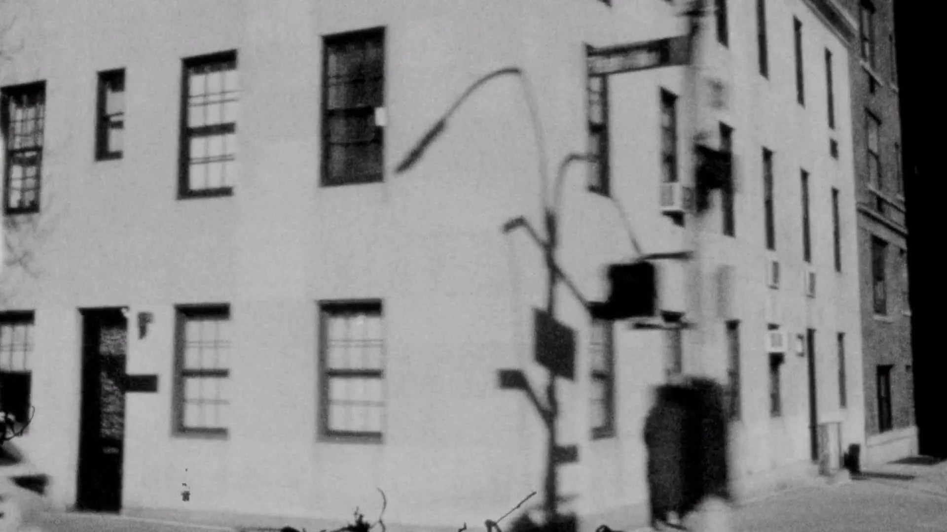 A black and white, grainy, shaky shot of a street corner with a building, a street sign, and a person walking, with a car passing by.