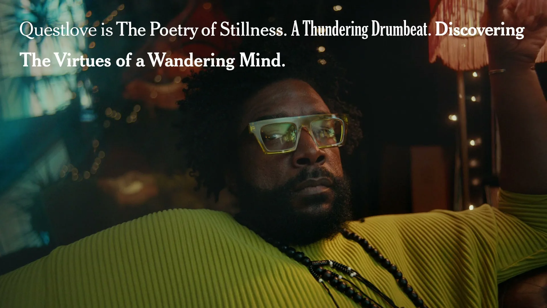 Questlove, a Black man with a beard and glasses, looks directly at the camera while sitting in a dimly lit room with string lights.