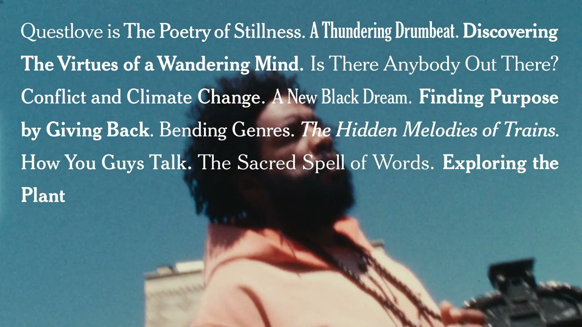 Questlove, a Black man with an afro and beard, wearing a peach hoodie and beaded necklaces, looks up as text appears on a blue sky background in an urban setting.