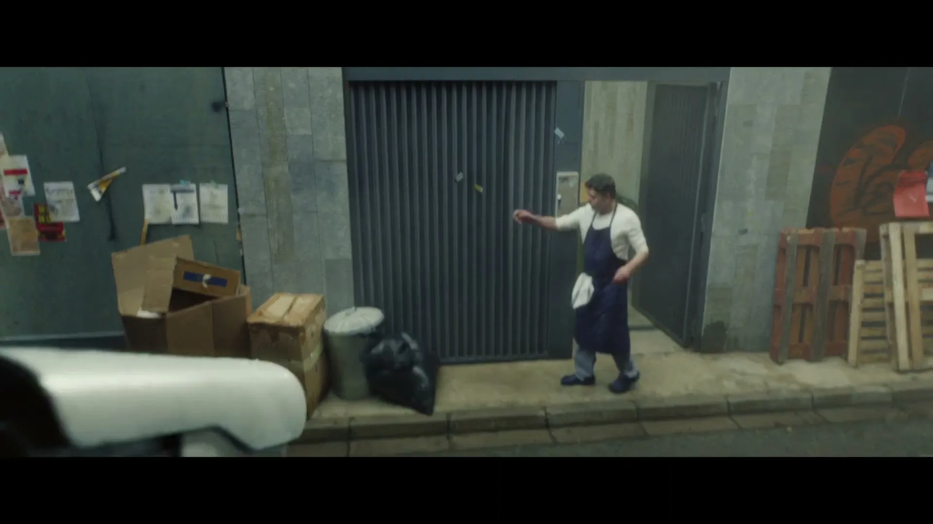 A male chef in an apron takes out the trash in a back alley, then turns to look at something off-screen.