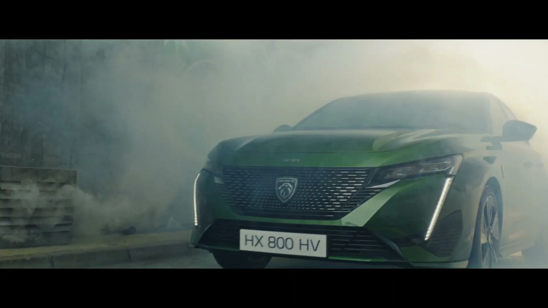 A green Peugeot car emerges from a cloud of smoke on a street, showcasing its front grille and headlights.