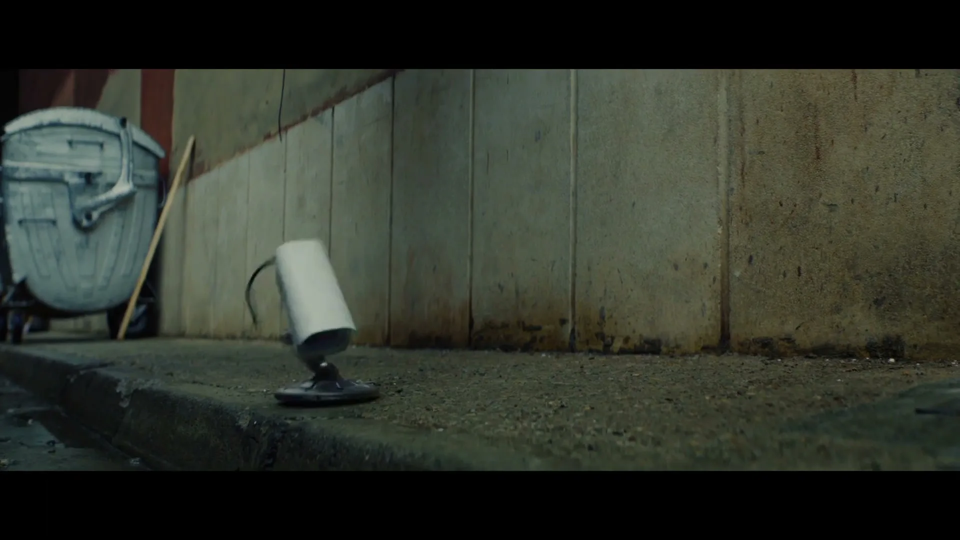 A small, white security camera with a green light detaches from its base and floats away from a dirty wall next to a trash can.