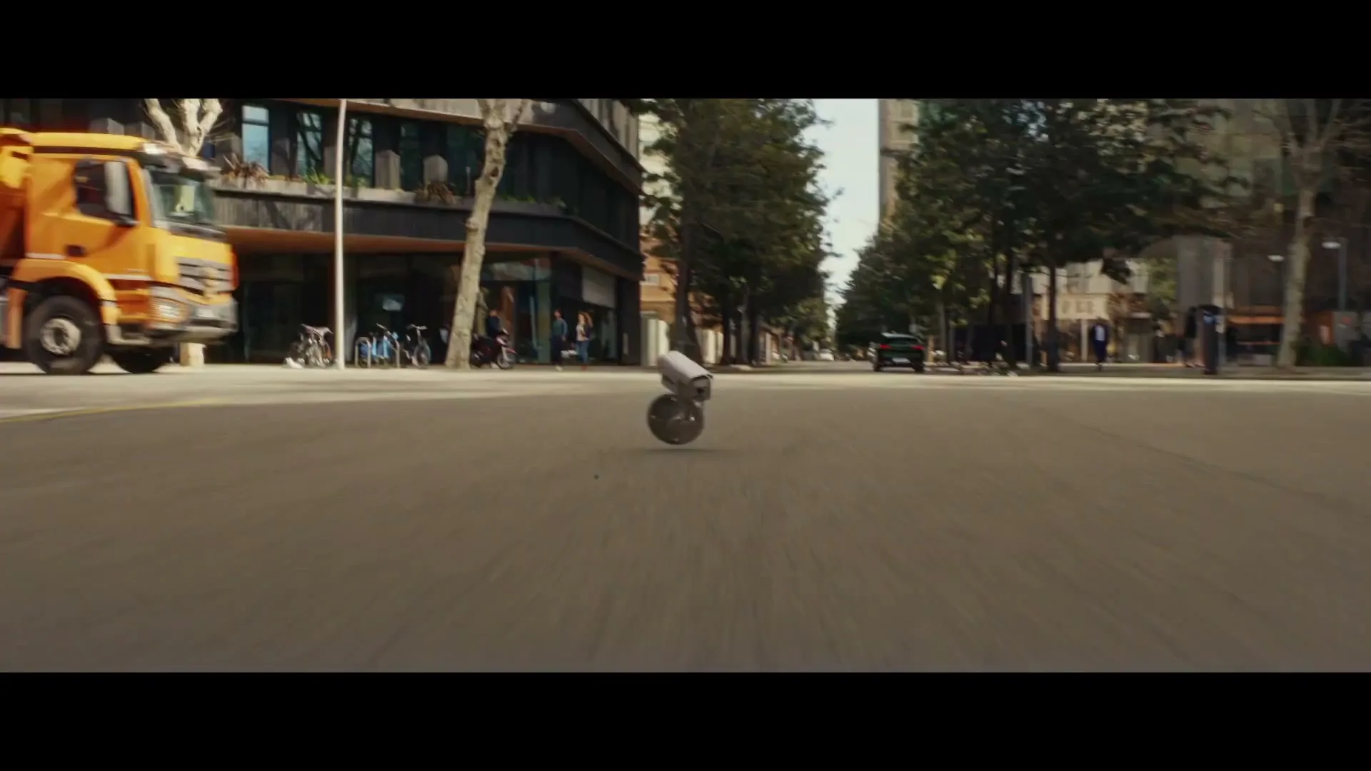A green Peugeot car drives down a city street, followed by a small camera on wheels that is nearly run over by a large orange truck.