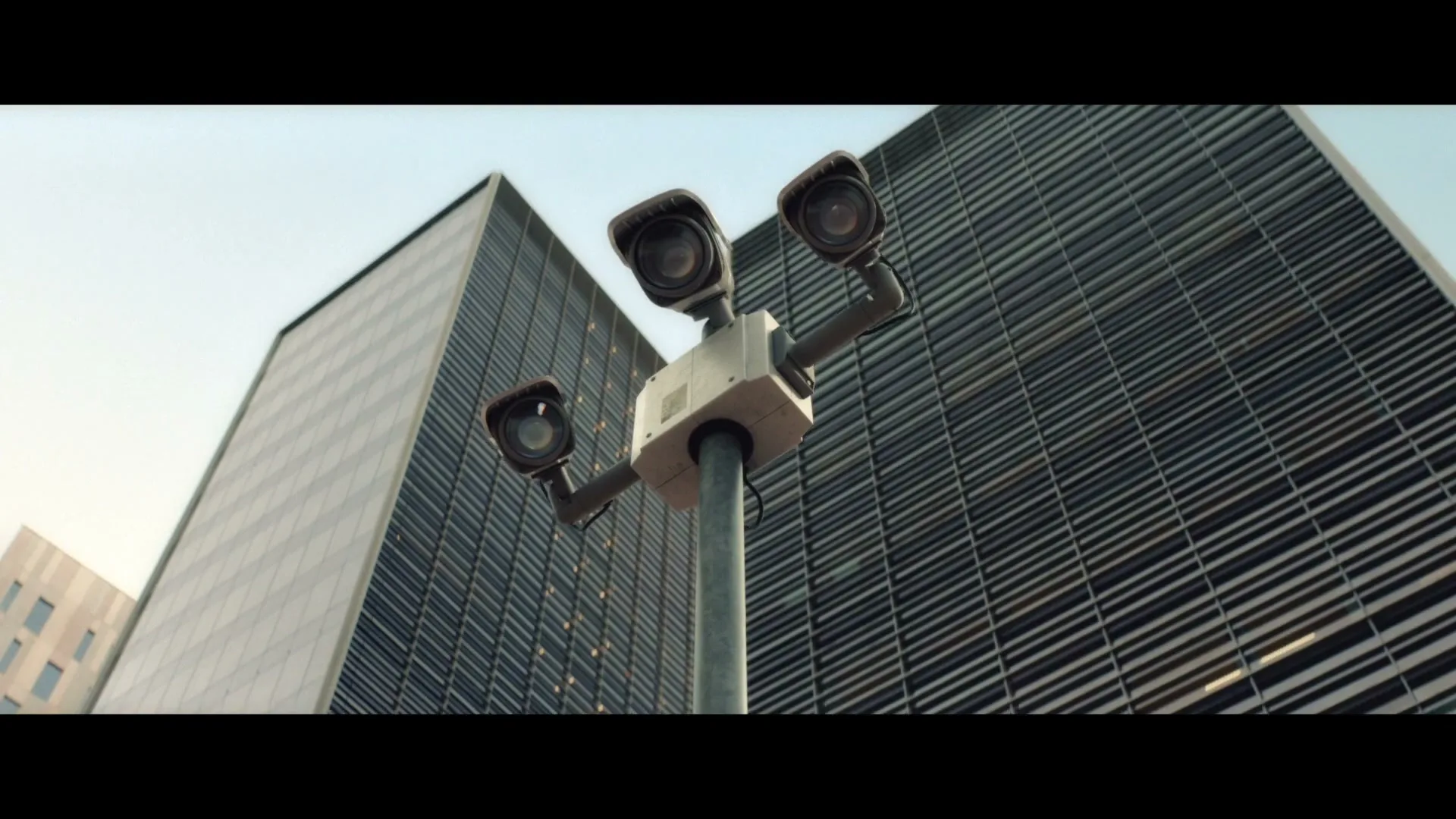 Three security cameras on a pole rotate, surveilling the area in front of tall, modern buildings under a clear sky.