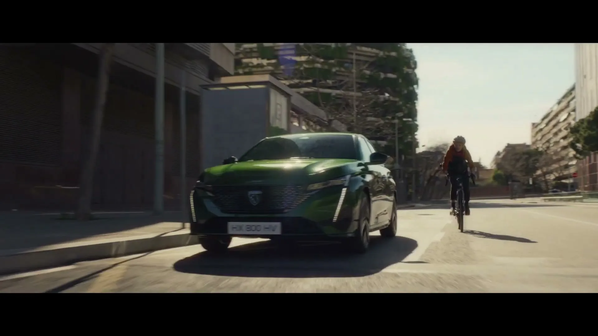 A green Peugeot car drives down a city street, with a cyclist riding alongside it and a pedestrian walking on the sidewalk.