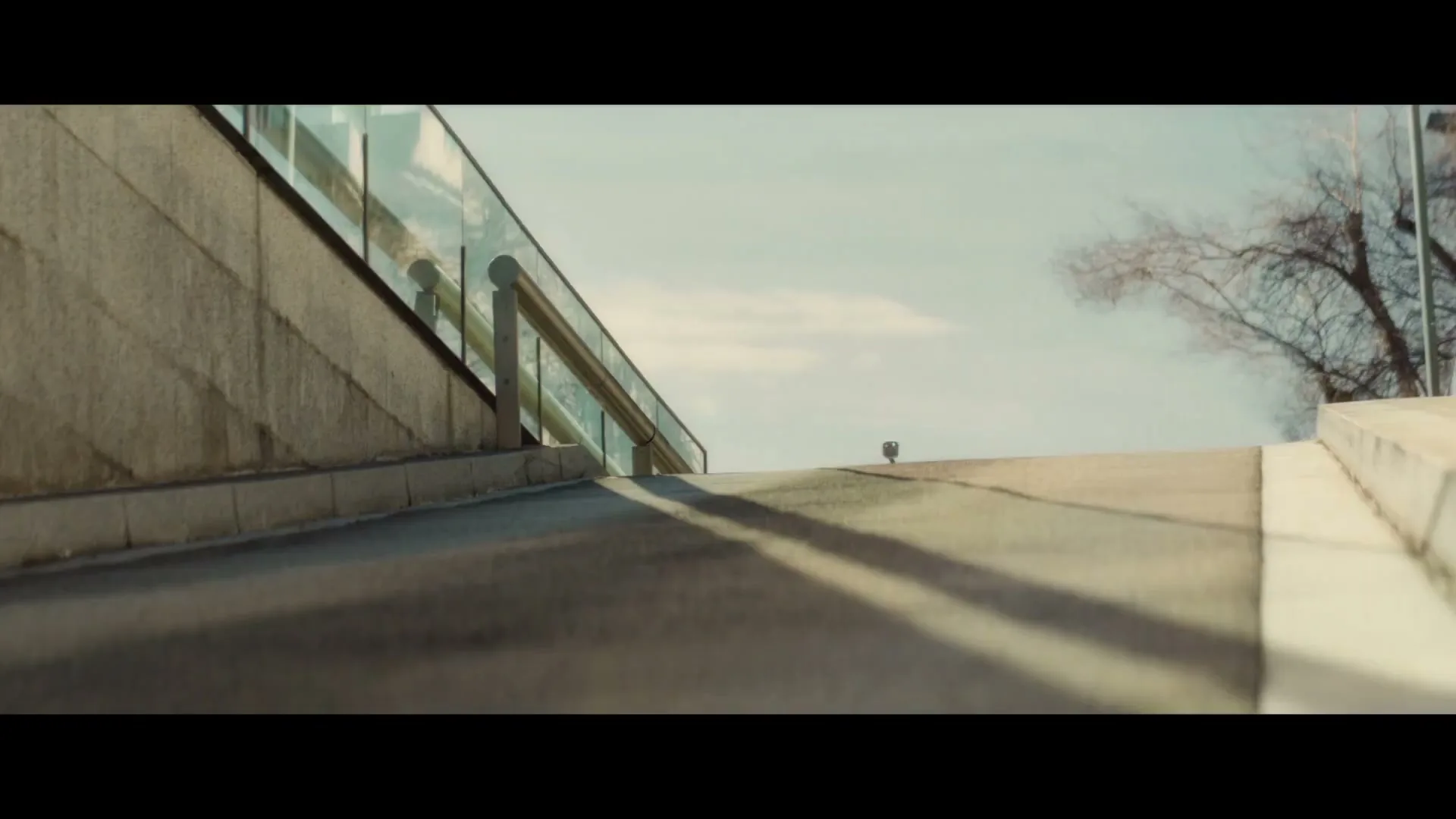 A low-angle shot of a concrete ramp leading up to a bridge with a glass railing, under a clear sky.