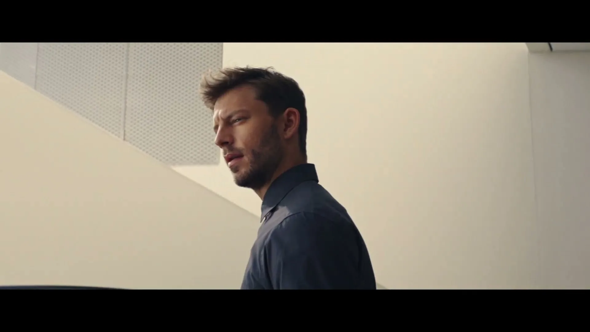 A man in his 30s, wearing a dark blue shirt, looks up and to the left with a curious expression in a modern, minimalist interior.
