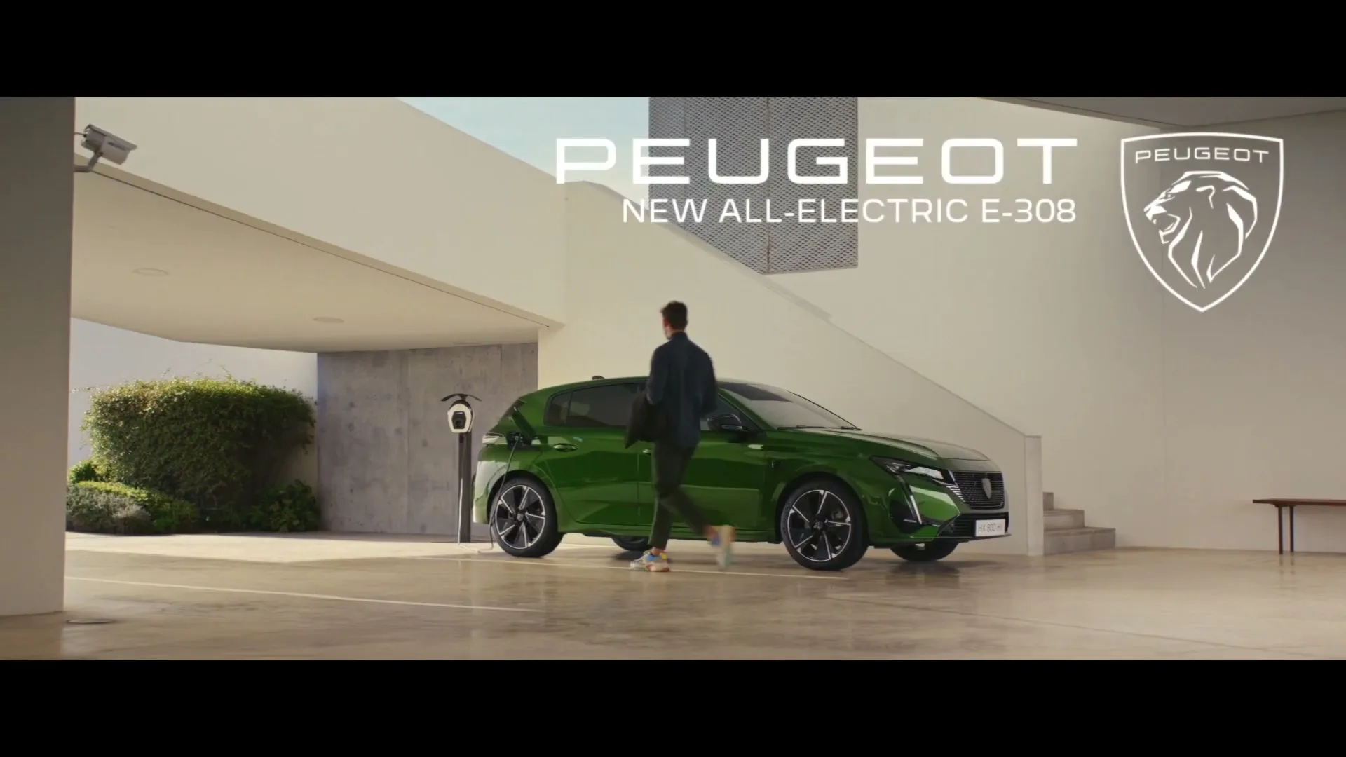 A man in a dark jacket and light pants walks towards a green Peugeot E-308 car, which is charging in a modern, open garage.