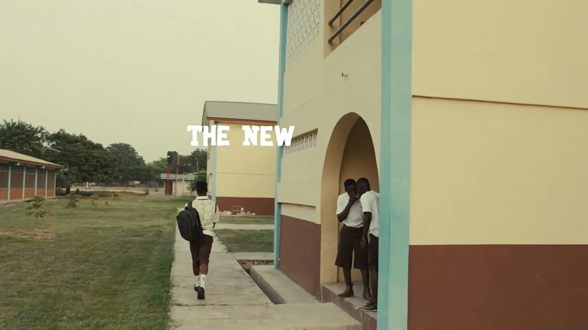 A young male student with a backpack walks away from the camera on a path next to a school building, while other students stand in a doorway.