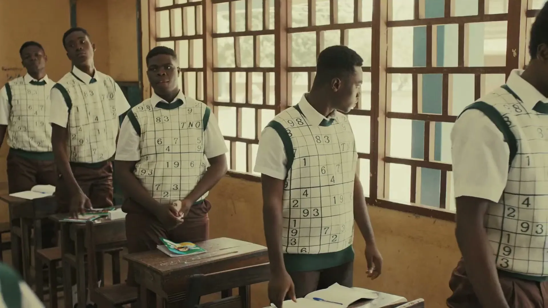 A group of male students in sudoku-patterned vests stand in a classroom, looking around with curious expressions.
