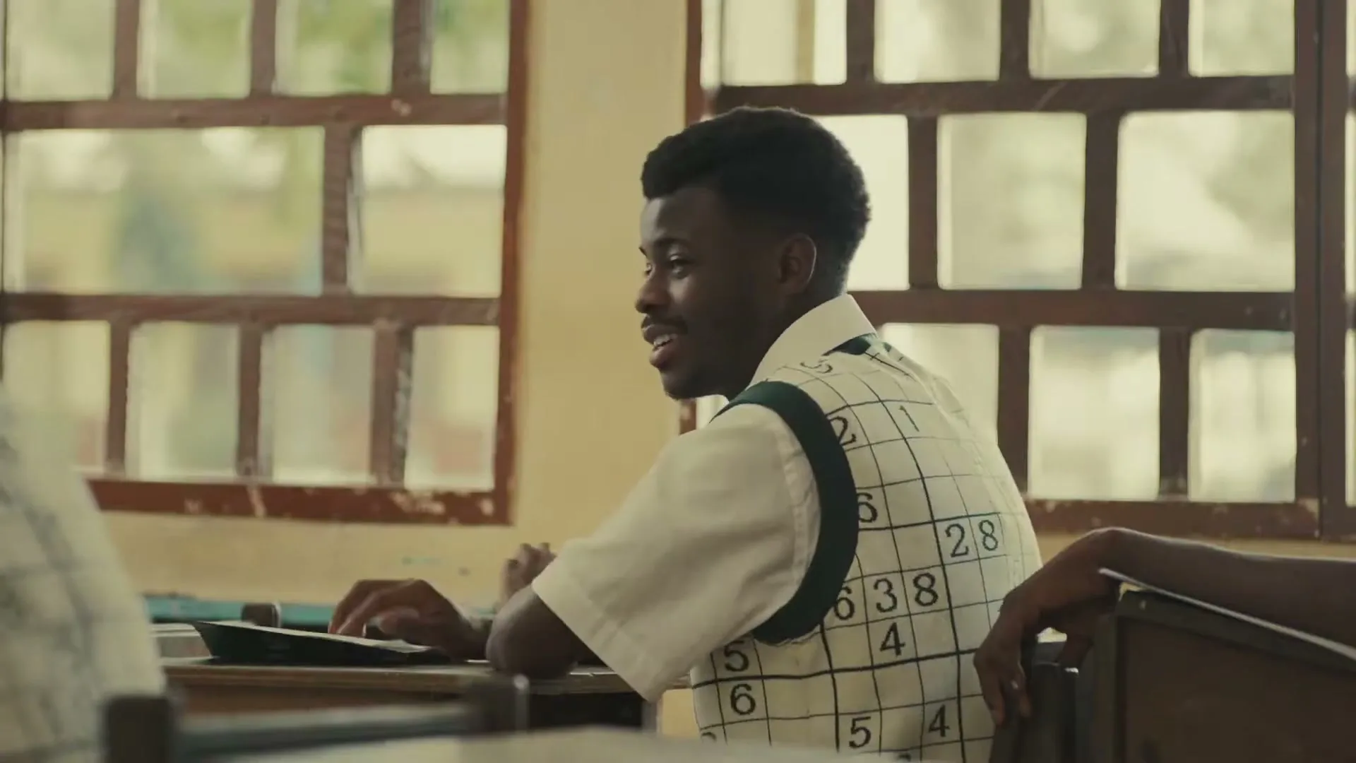 A young Black man in a sudoku vest sits at a desk in a classroom, looking up and smiling.