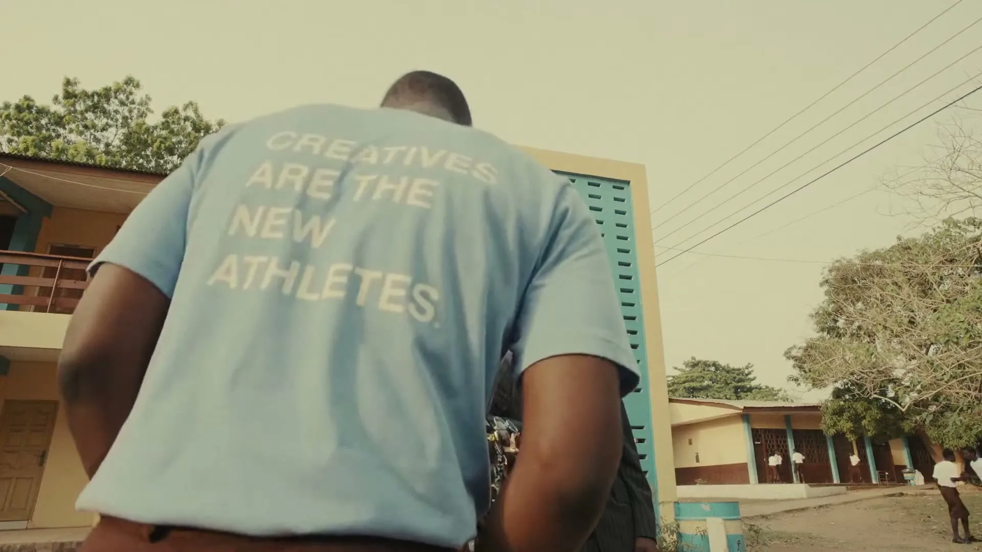 A young man in a light blue t-shirt with text on the back walks past another young man in a suit and sunglasses at a school.