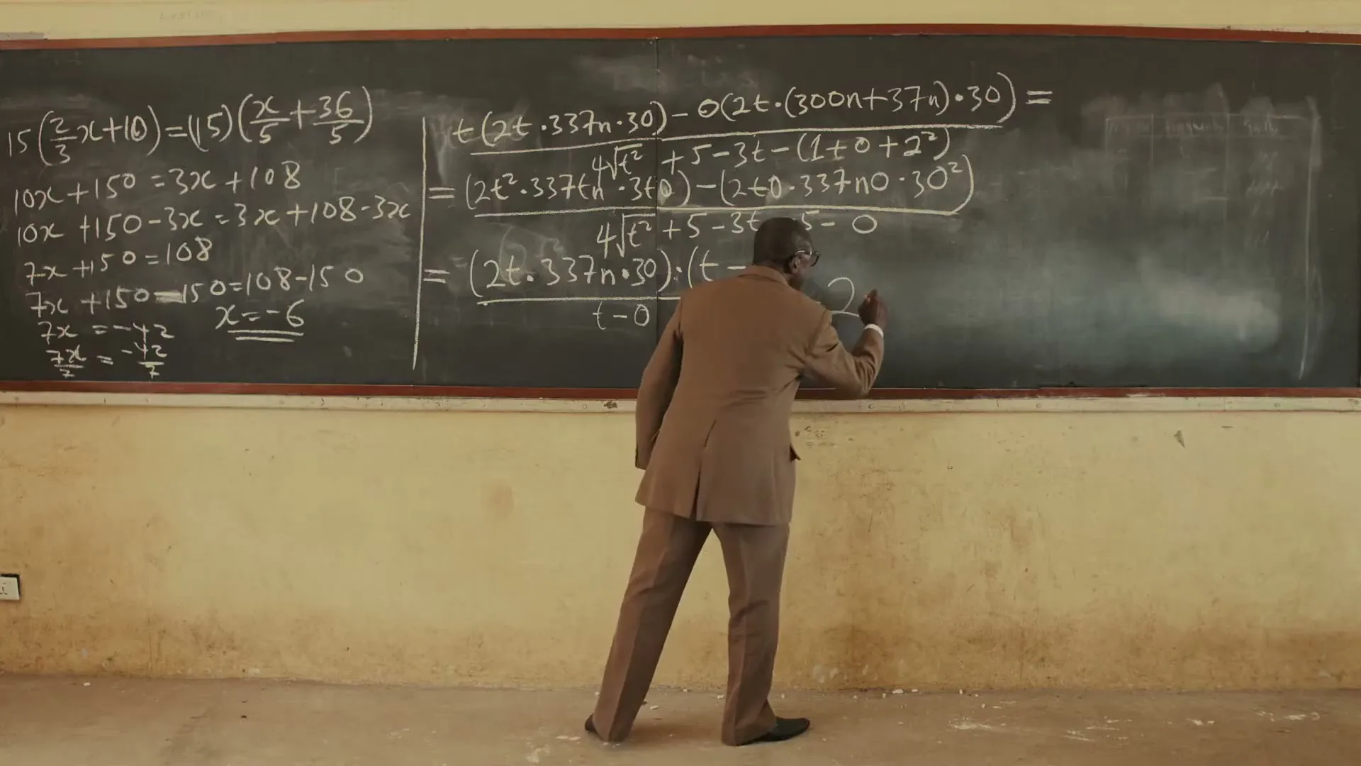 A male teacher in a brown suit writes complex math equations on a large blackboard in a classroom.