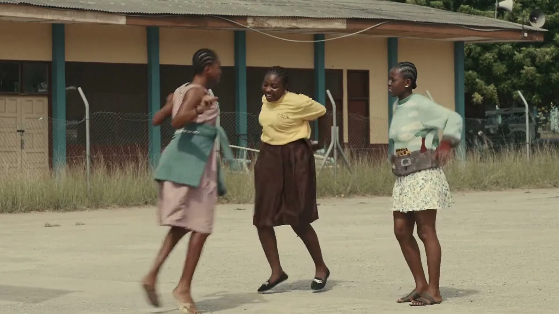 Three young women in casual clothes dance joyfully in an open, dusty area outside a building, with one woman in a yellow shirt and brown skirt, another in a striped dress and green apron, and a third in a light green sweater and floral skirt.