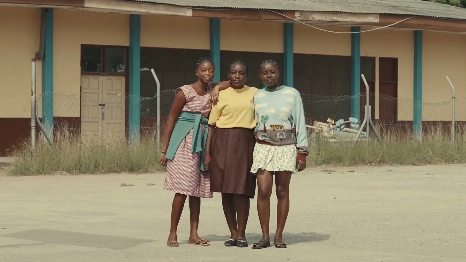 Three young Black women stand together, one with her arm around another, in front of a school building on a sunny day.