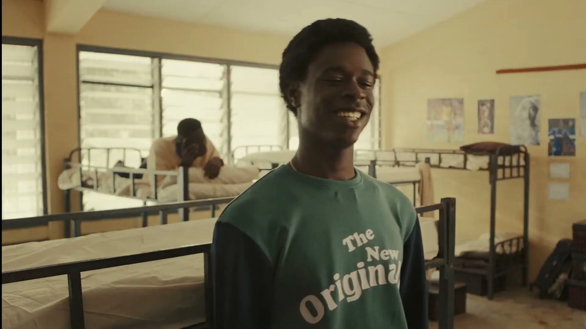 A young Black man in a green and blue sweatshirt smiles and looks up, standing in a dorm room with bunk beds.