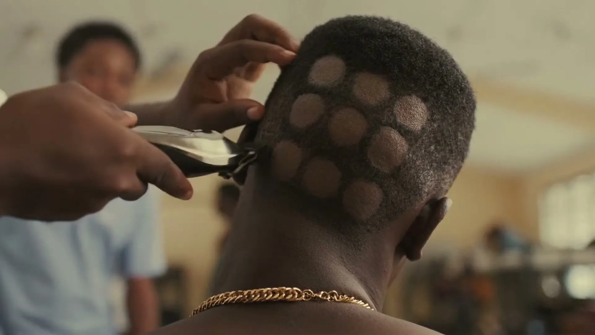 A barber shaves a pattern of circles into the back of a young man's head, who wears a gold chain, in a barbershop.