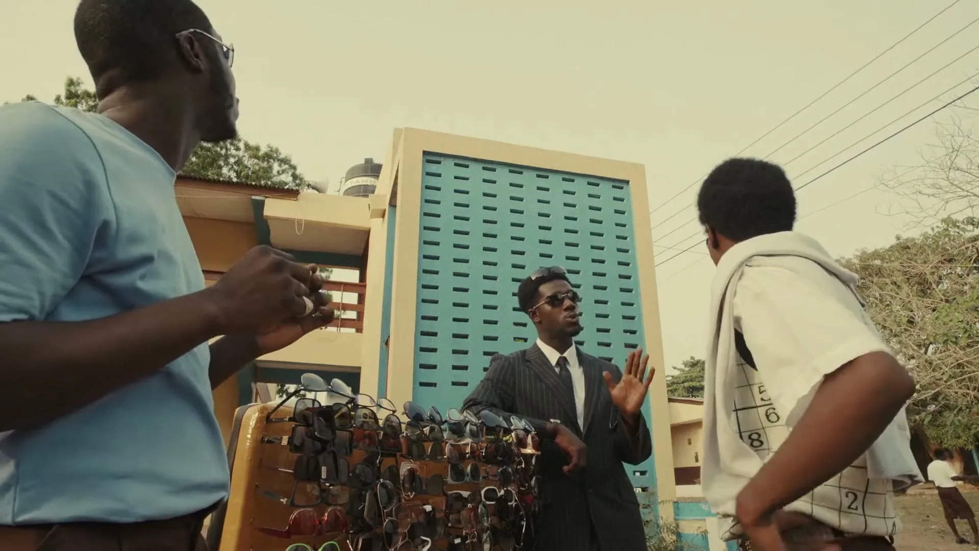 Three men are outside, two of them looking at sunglasses on a stand, while the third man in a suit talks to them, with a building in the background.