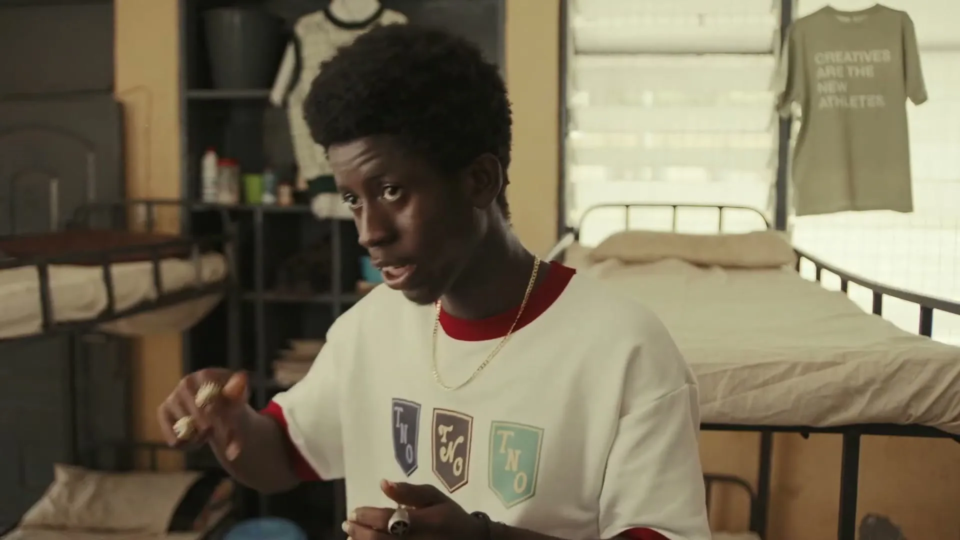 A young Black man with an afro, wearing a white t-shirt with colorful crests, talks and gestures in a sparse dorm room with bunk beds.