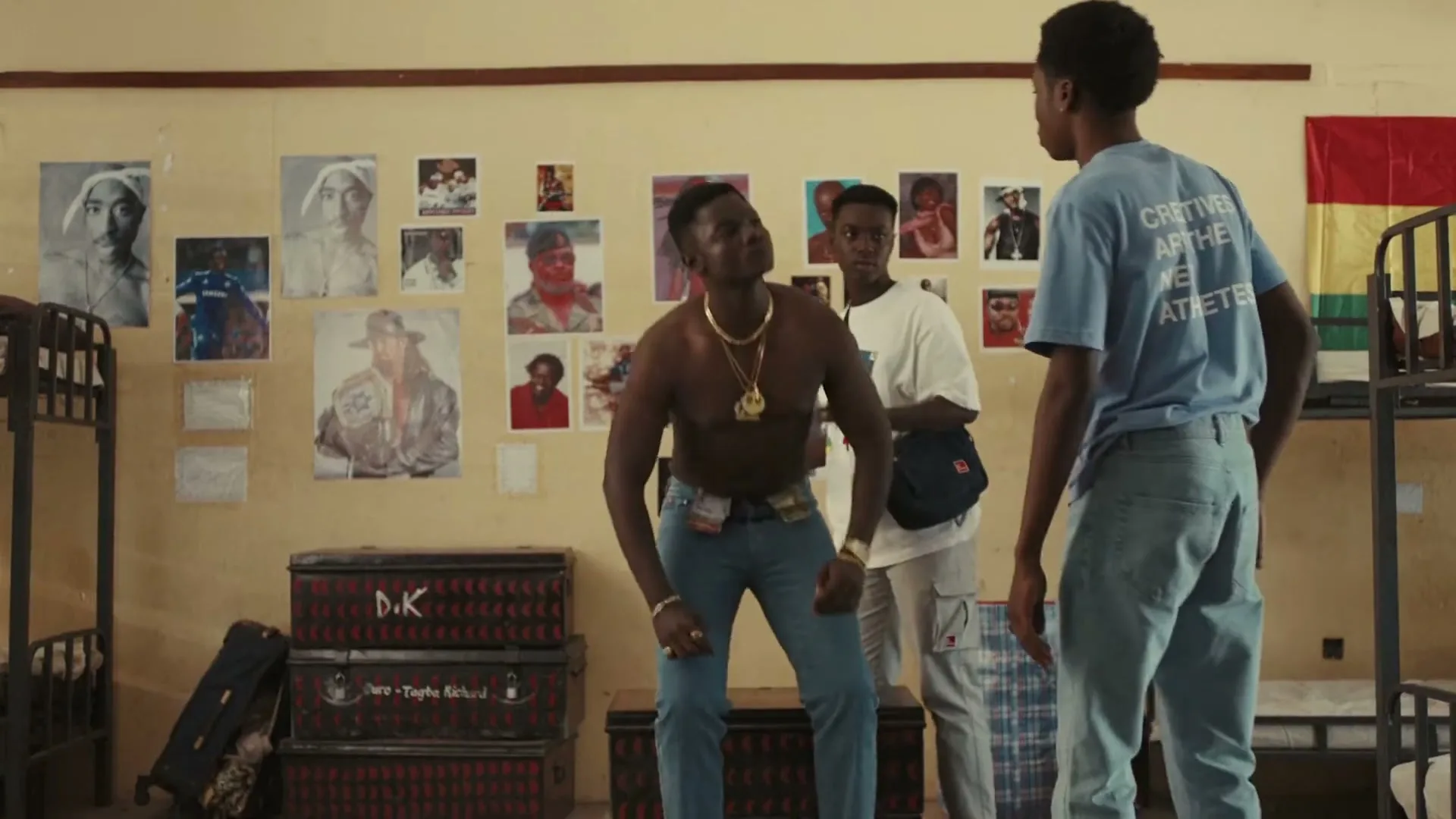 Three young men are in a dorm room, one shirtless and wearing gold chains, standing up from a trunk while another adjusts his hair.
