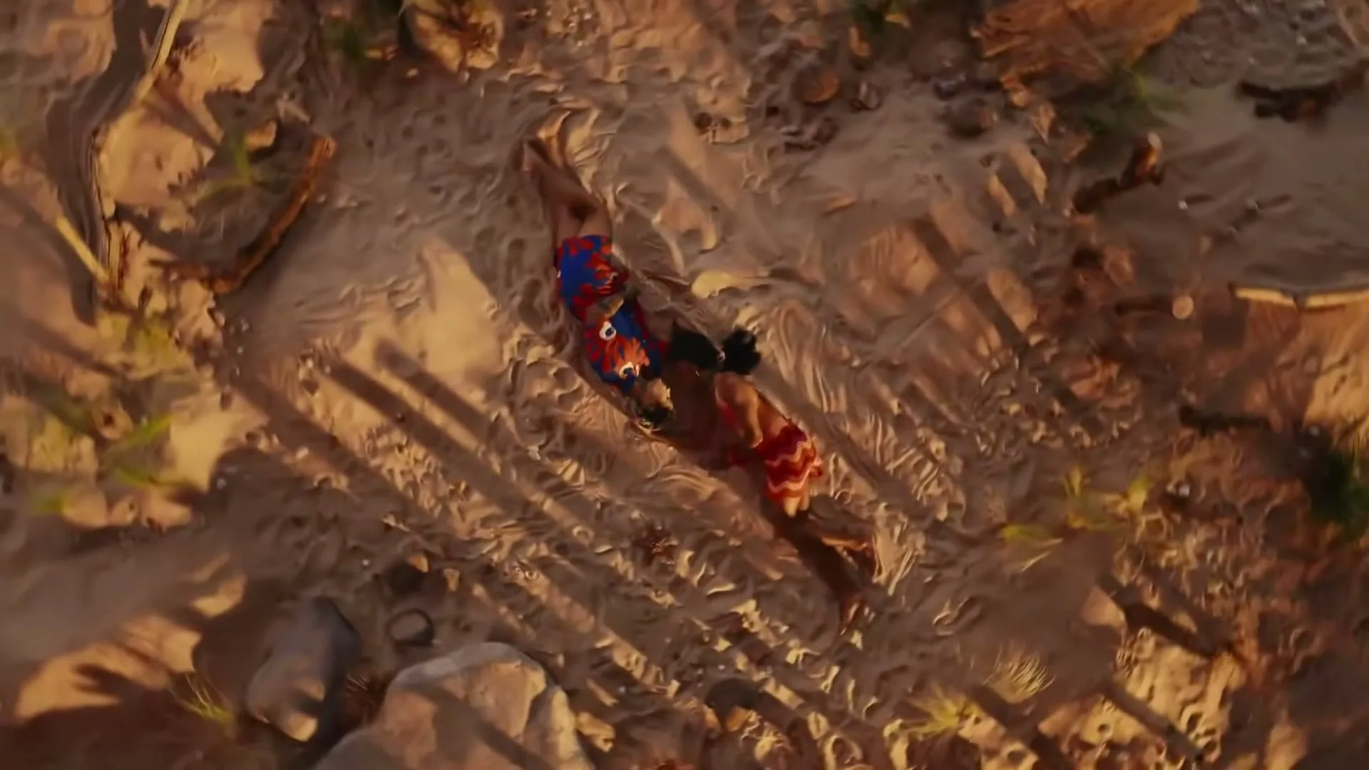 Two people in colorful swimsuits are lying on a sandy beach, seen from an overhead perspective, as the camera slowly zooms out.