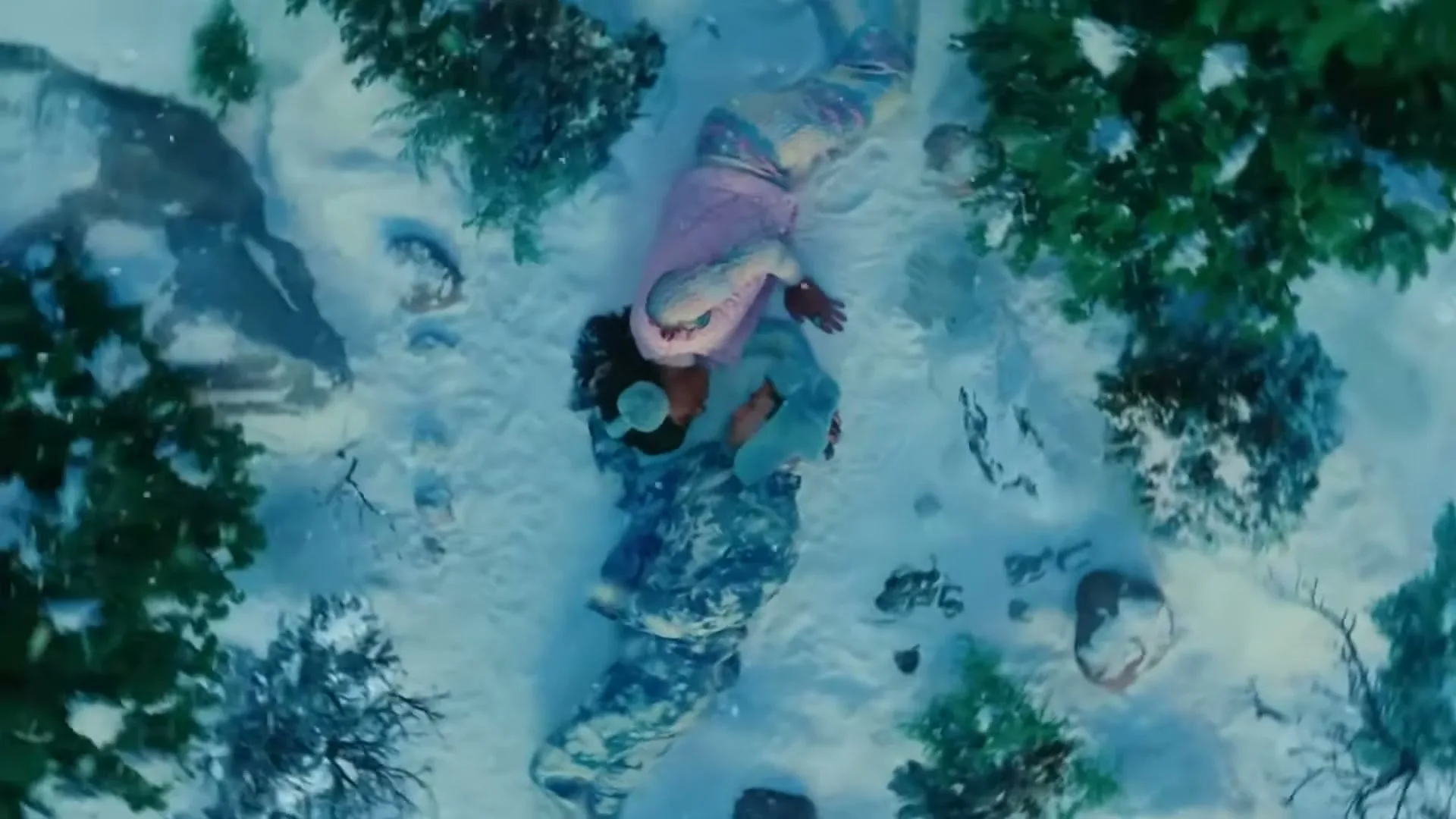 A couple in winter clothes lies on their backs in the snow, surrounded by small snowy trees, as the camera slowly zooms out.