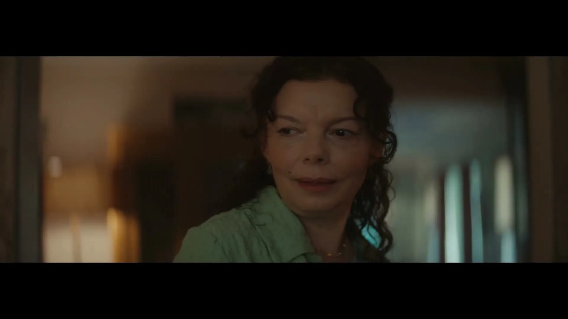 A woman with curly brown hair and a green shirt turns to the camera and smiles, then looks down, in a dimly lit room.