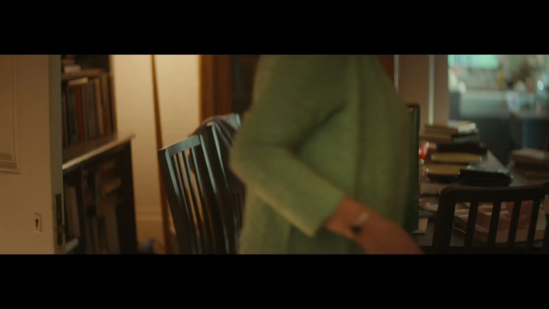 A person in a green sweater places a green Uber Eats bag on a cluttered dining table in a home.