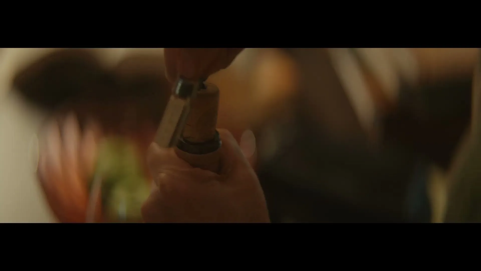 A person's hands use a corkscrew to remove a cork from a wine bottle in a dimly lit interior.