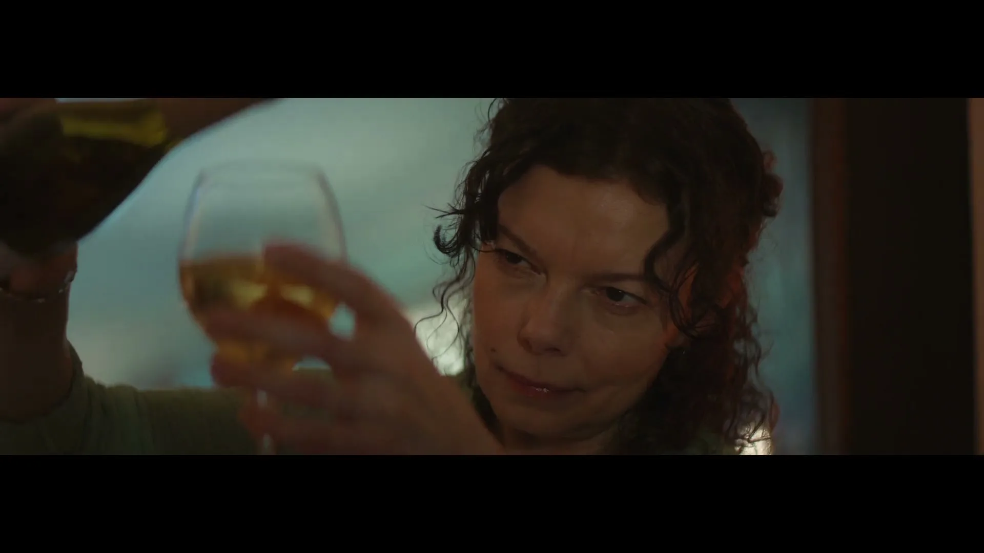 A woman with curly brown hair pours white wine into a glass, looking intently at the liquid as it fills the glass in an interior setting.