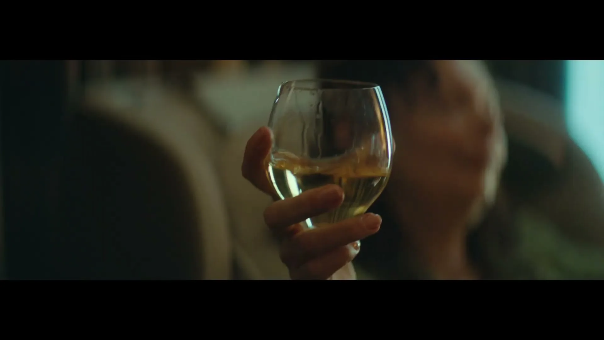A woman's hand holds a glass of white wine, swirling it gently, in a dimly lit interior setting.