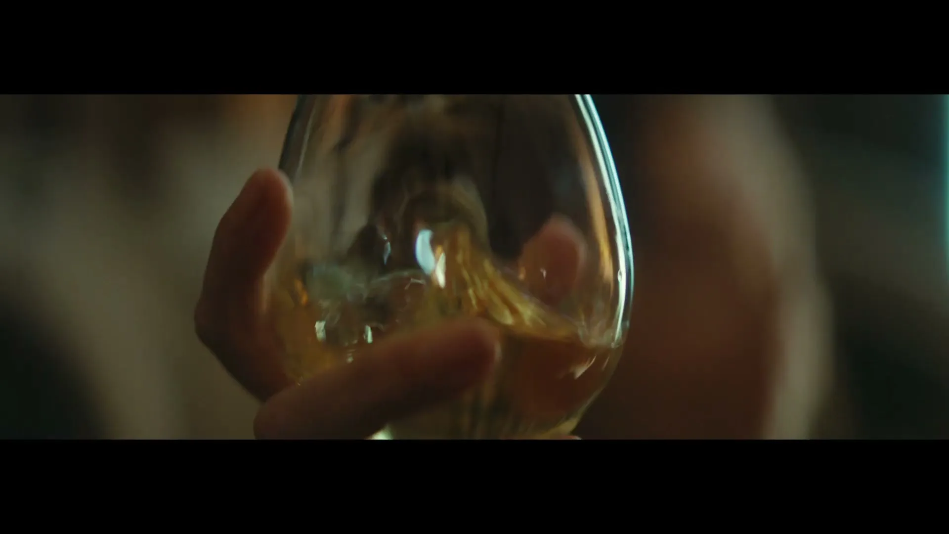 A person's hand holds a glass of amber liquid, swirling it gently, in a dimly lit interior setting.