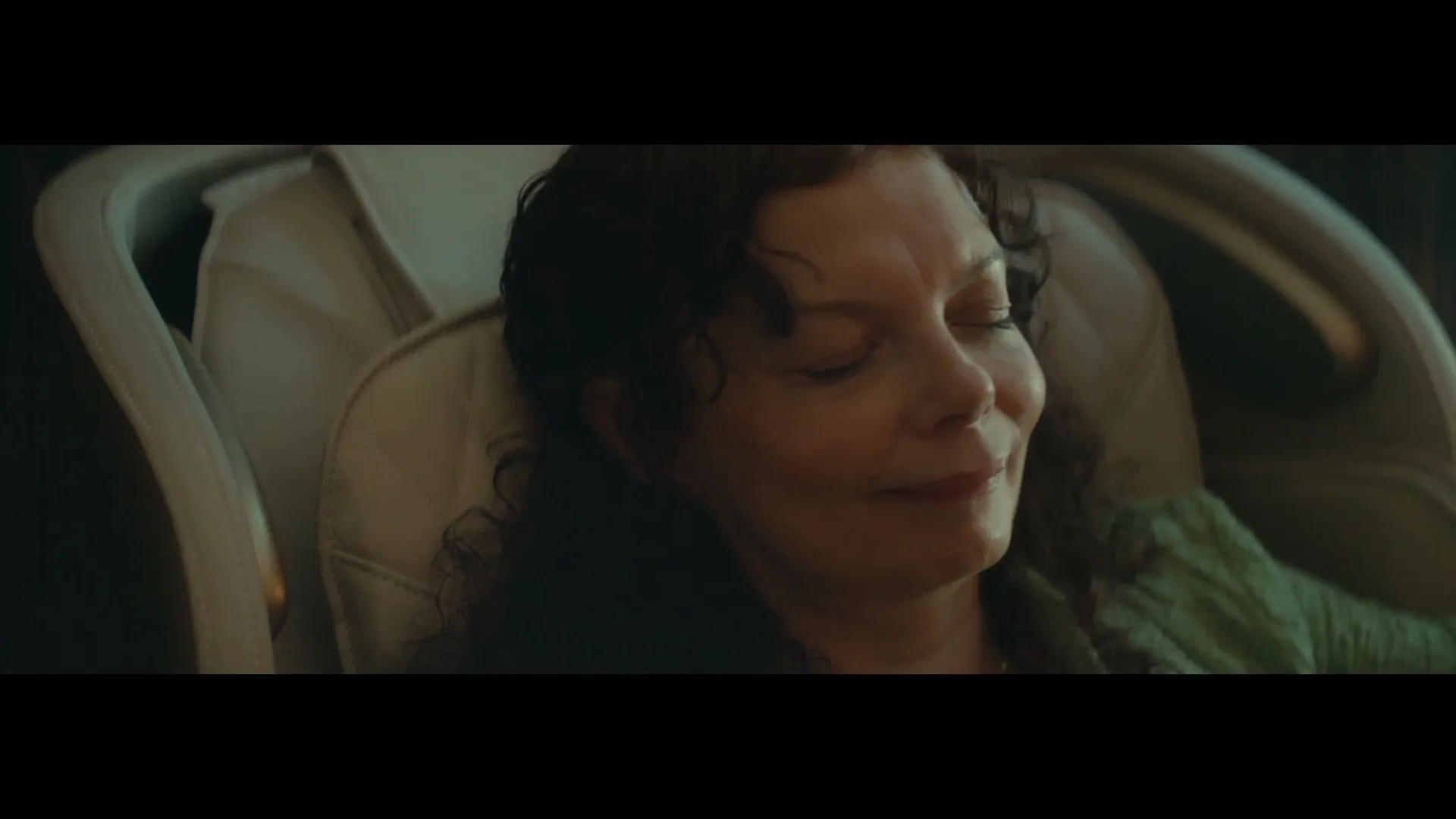 A woman with dark curly hair and a green top smiles with her eyes closed while sitting in a light-colored massage chair.