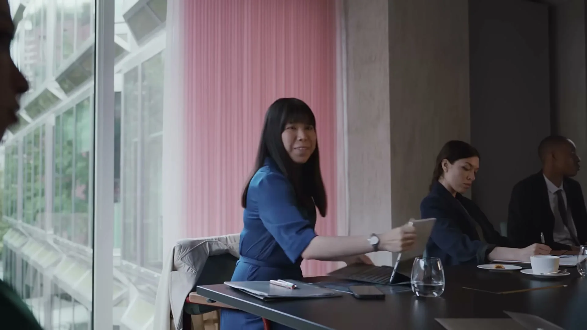 A young Asian woman in a blue dress closes her laptop at a conference table, with other colleagues visible in a modern office setting.