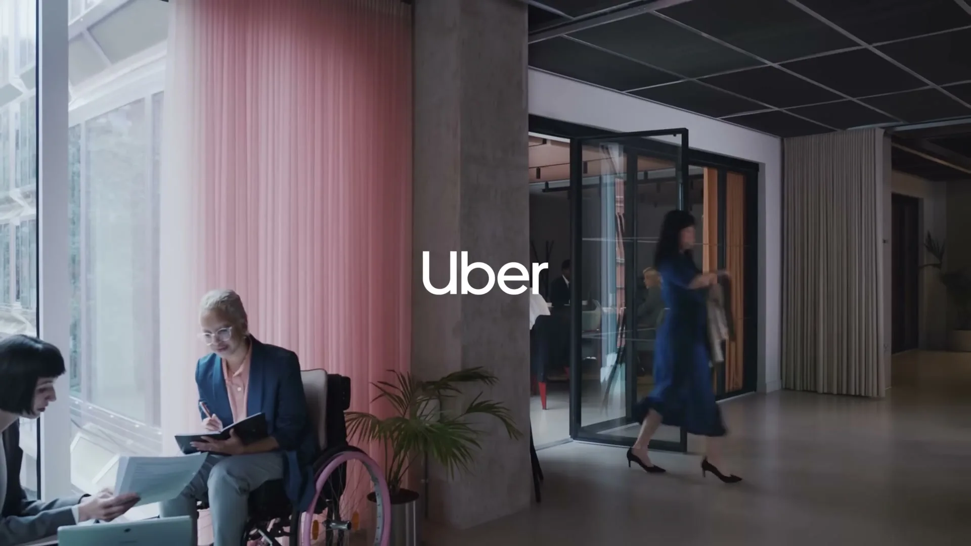 A woman in a wheelchair and another woman are talking by a window, while a third woman walks past them in an office.