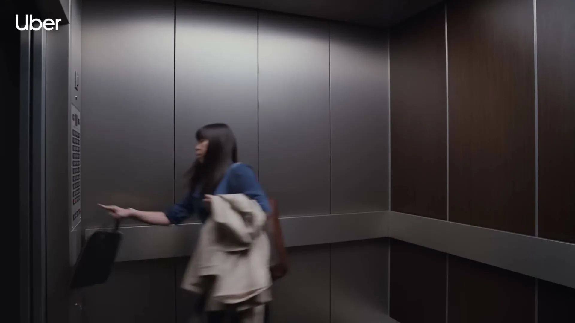 A young woman in a blue dress and carrying a coat enters an empty elevator and presses a button, then looks up in surprise.