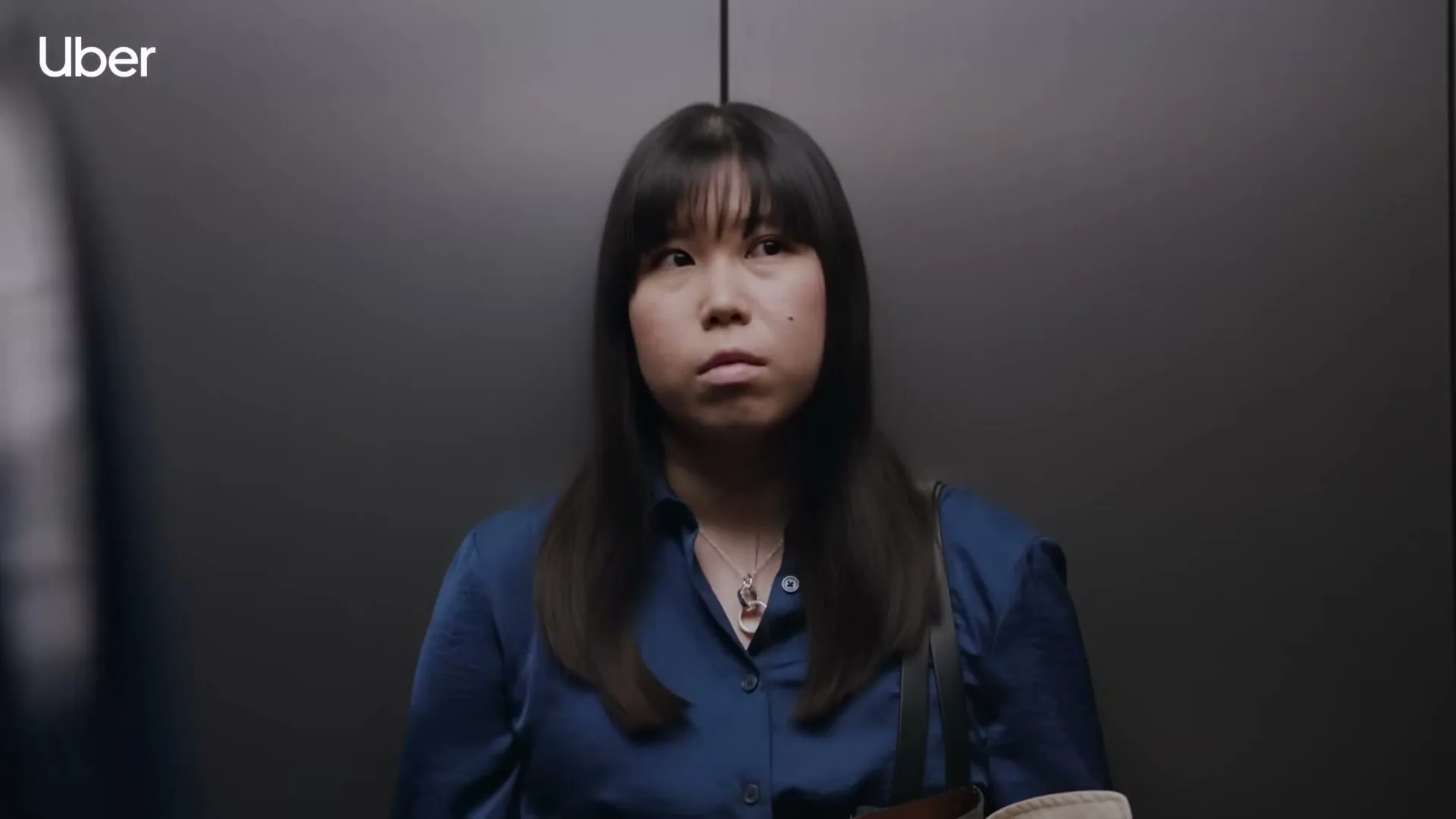 A young Asian woman with long dark hair and a blue shirt stands in an elevator, looking slightly annoyed as a man's shoulder partially obstructs the view.
