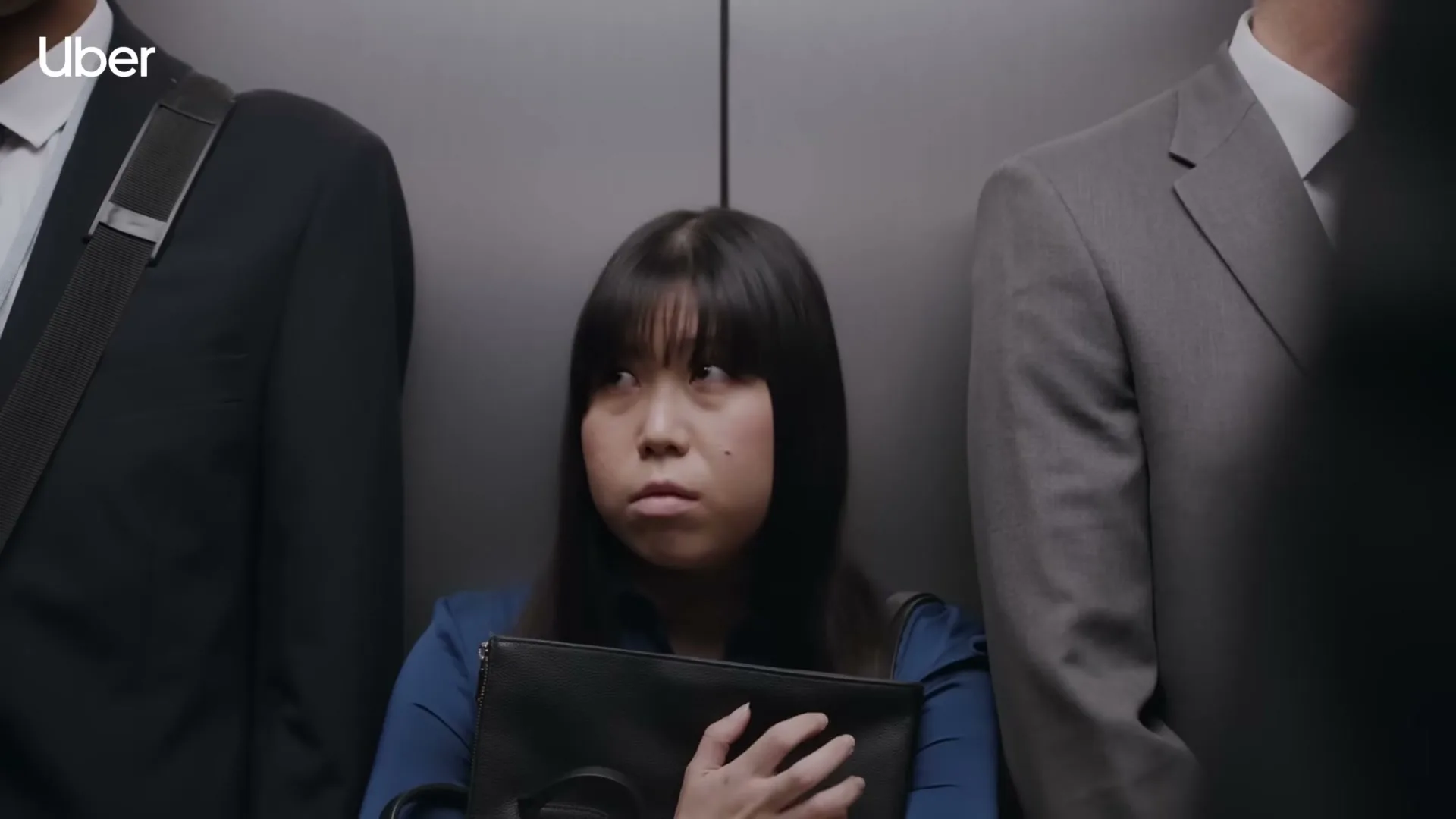 A young Asian woman in a blue shirt stands uncomfortably in a crowded elevator, surrounded by men in suits.
