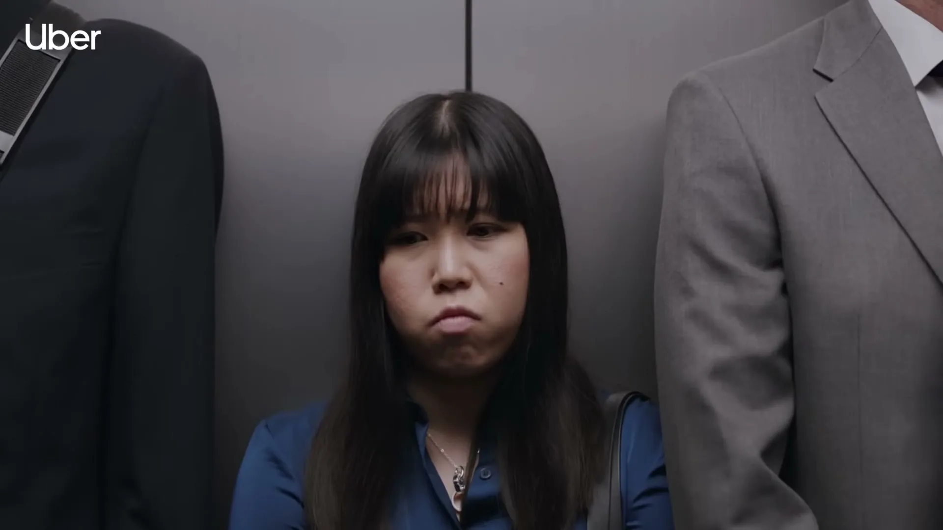 A young Asian woman with long dark hair and a blue shirt looks annoyed while standing in a crowded elevator between two men in suits.