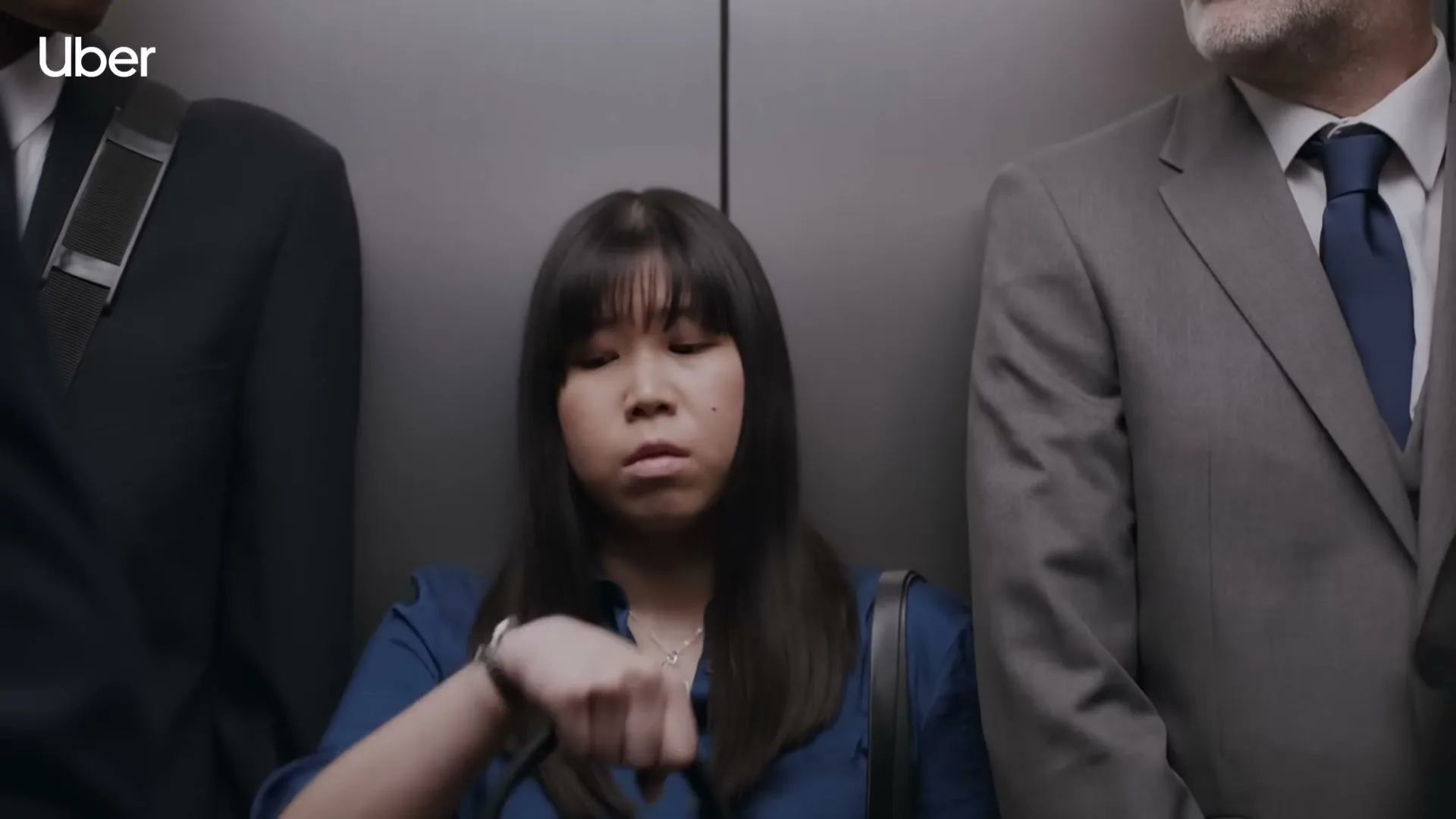 A young Asian woman in a blue shirt checks her watch anxiously while standing in a crowded elevator between two men in suits.