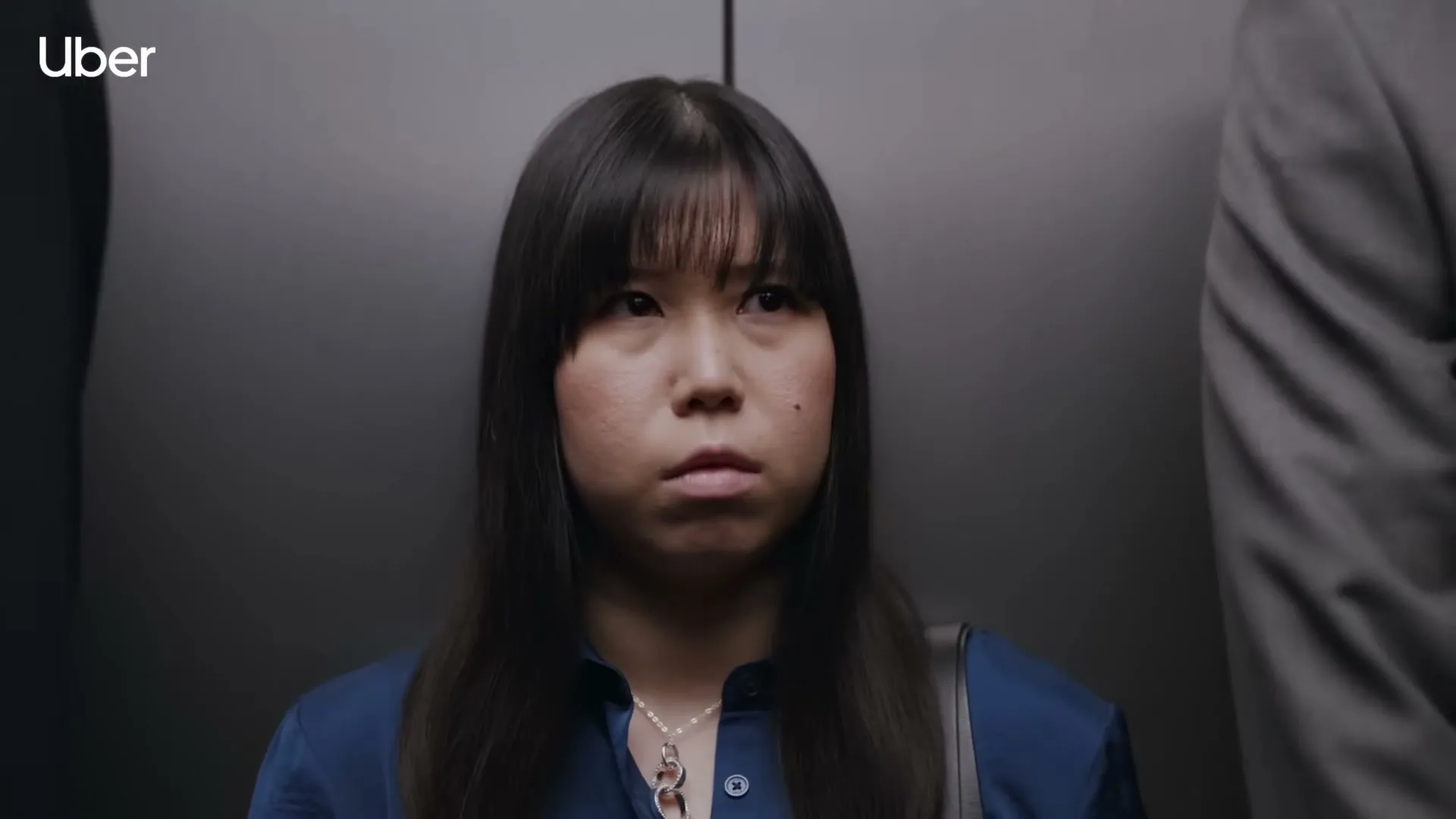 A young Asian woman with long dark hair and bangs stands in an elevator, looking around with a worried expression.