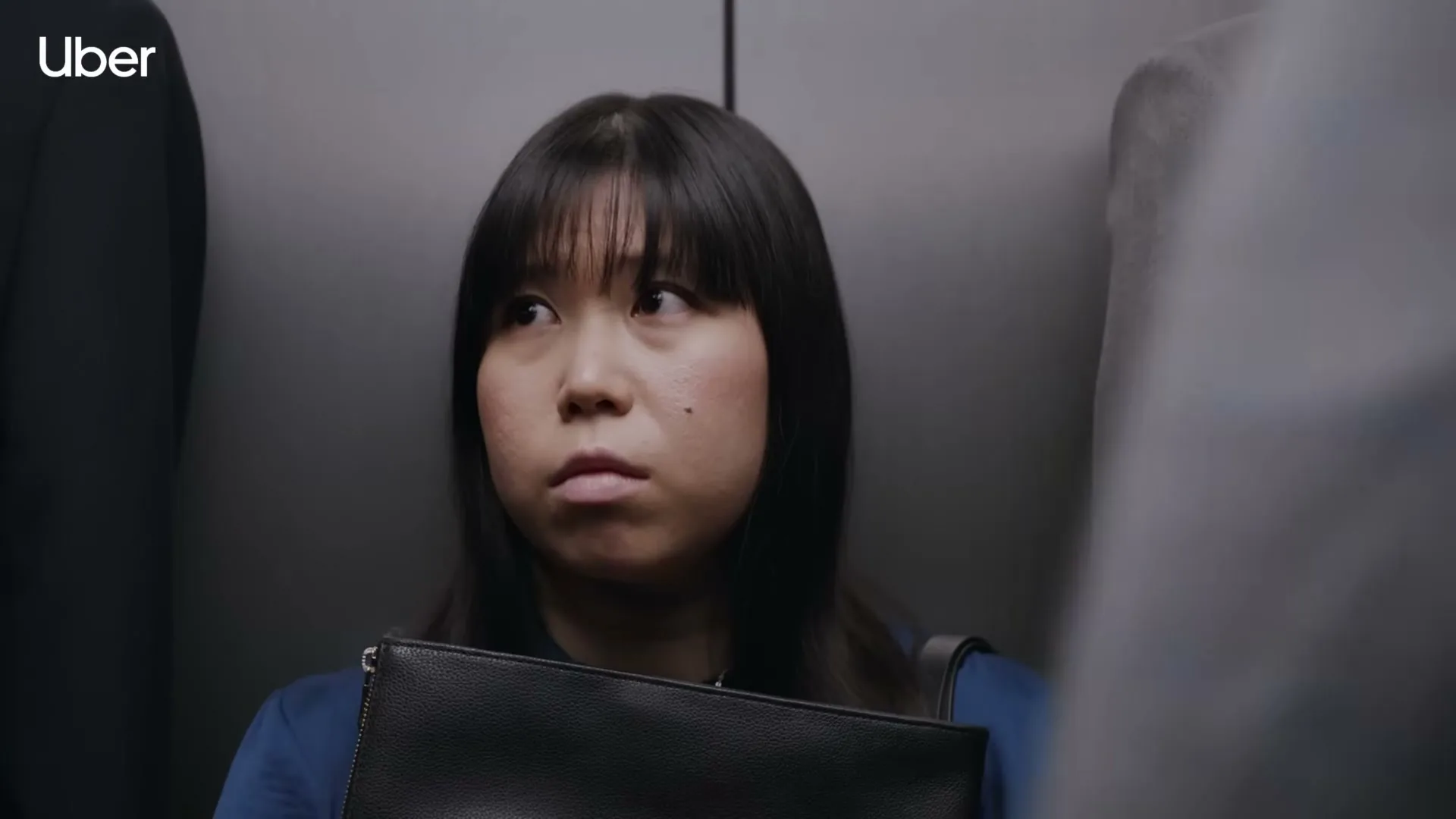 A young Asian woman with bangs and a blue top looks around nervously in a crowded elevator, holding a black clutch.