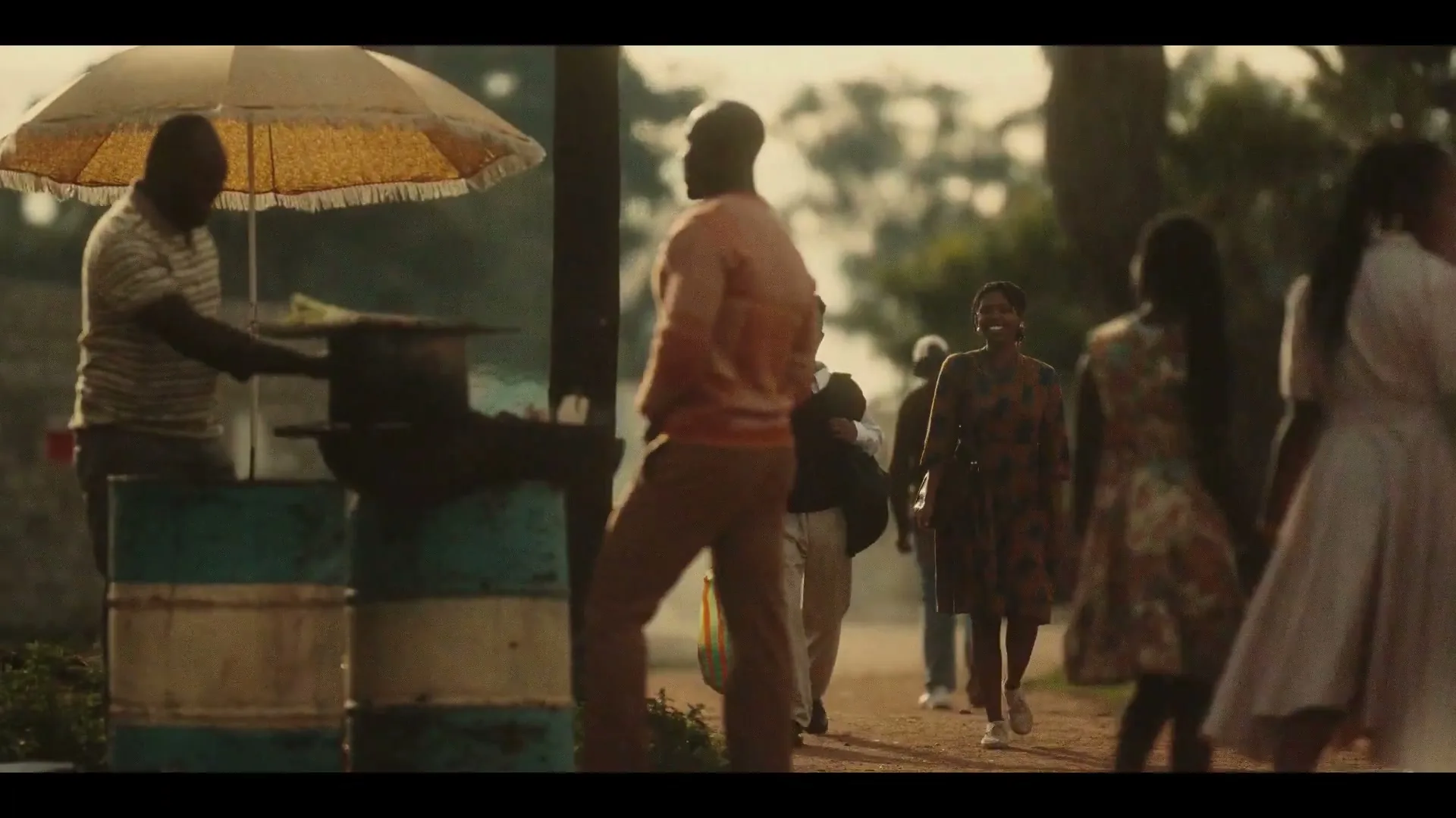 A diverse group of people walk along a dirt path, some smiling, past a street vendor under a yellow umbrella, in a sun-drenched outdoor setting.
