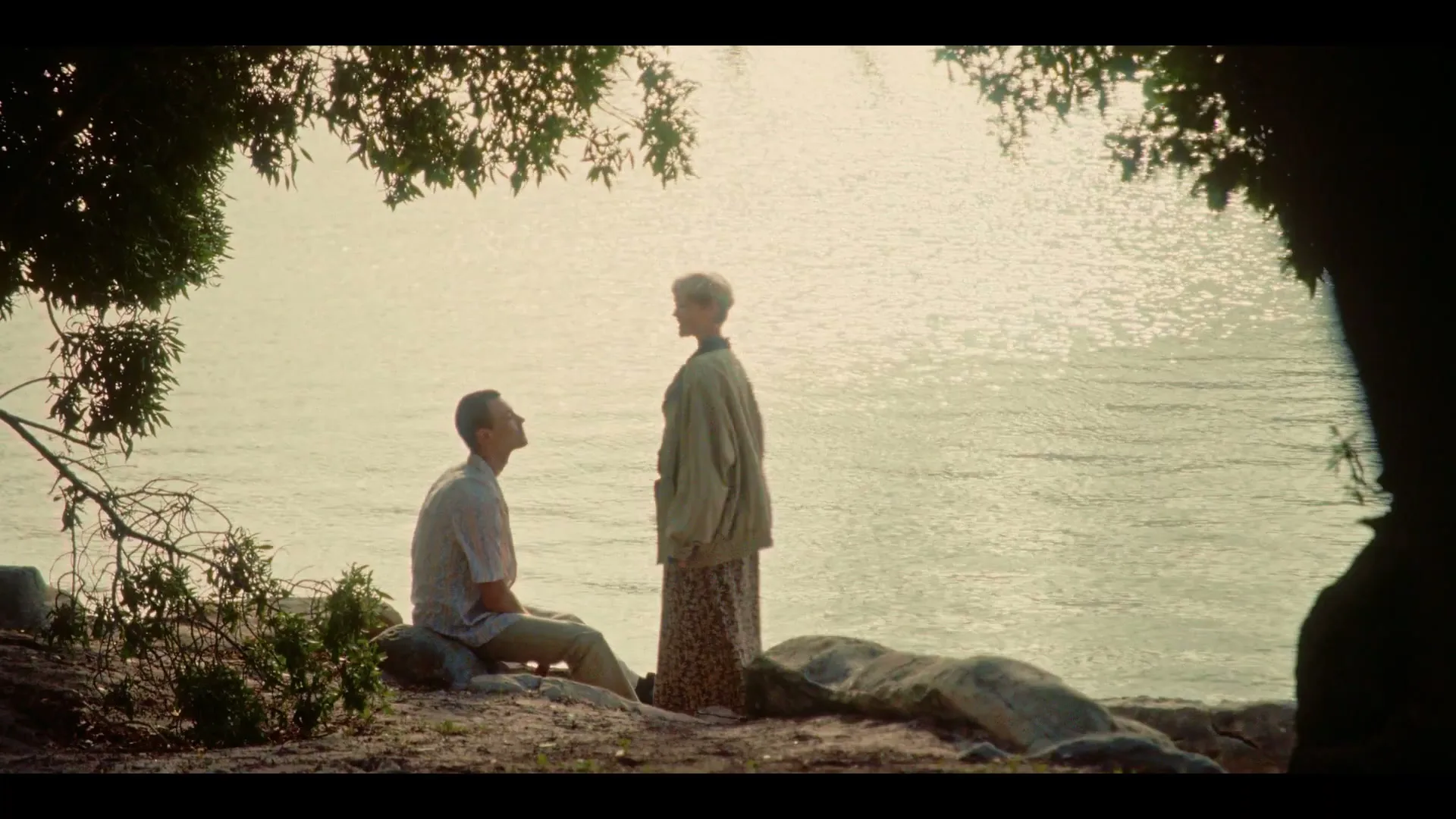 A woman with short blonde hair stands over a man sitting on rocks by a lake, gently touching his face under a tree.