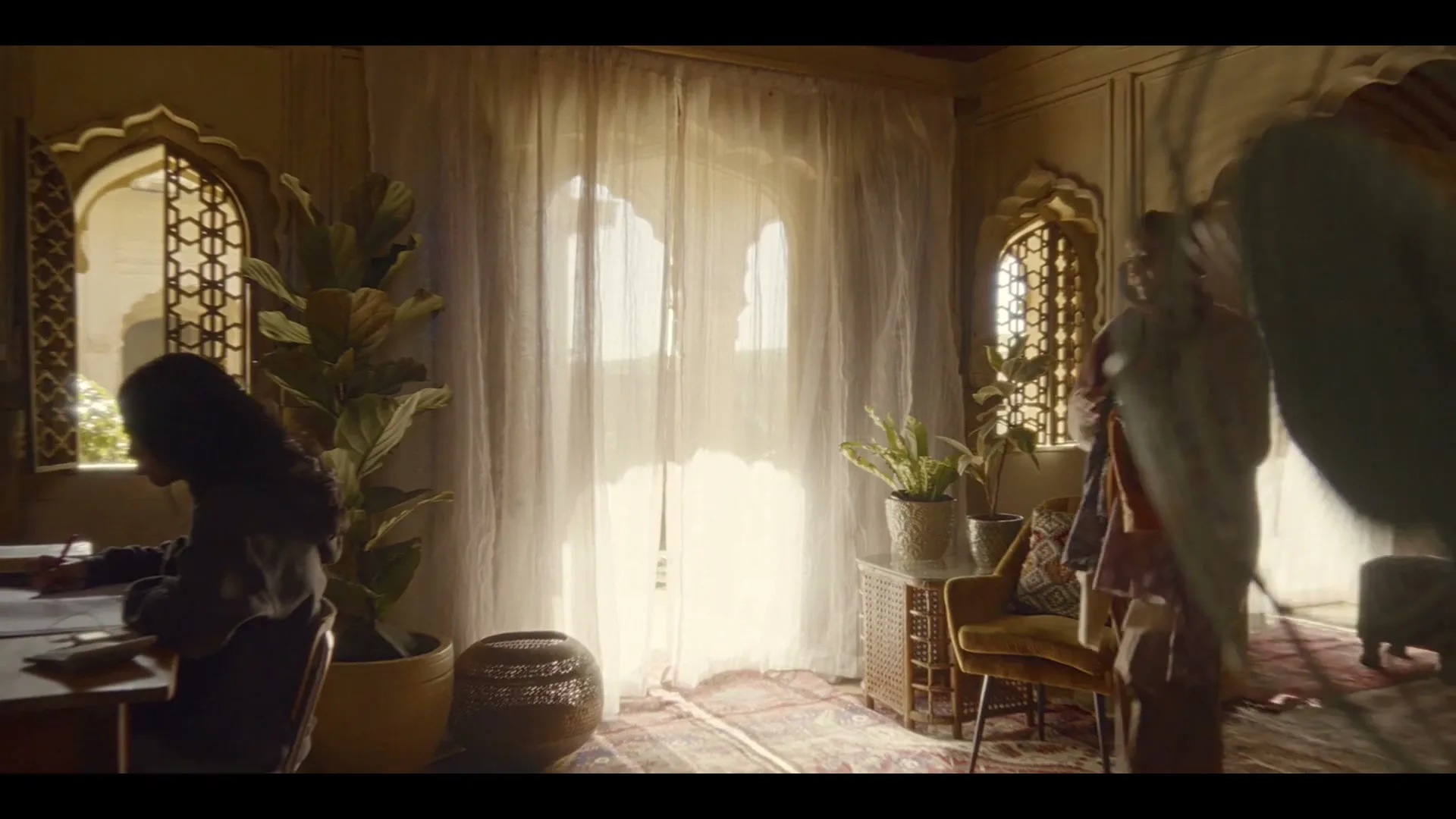 A young girl sits at a desk drawing, while an adult woman walks past her in a sunlit room with arched windows and sheer curtains.