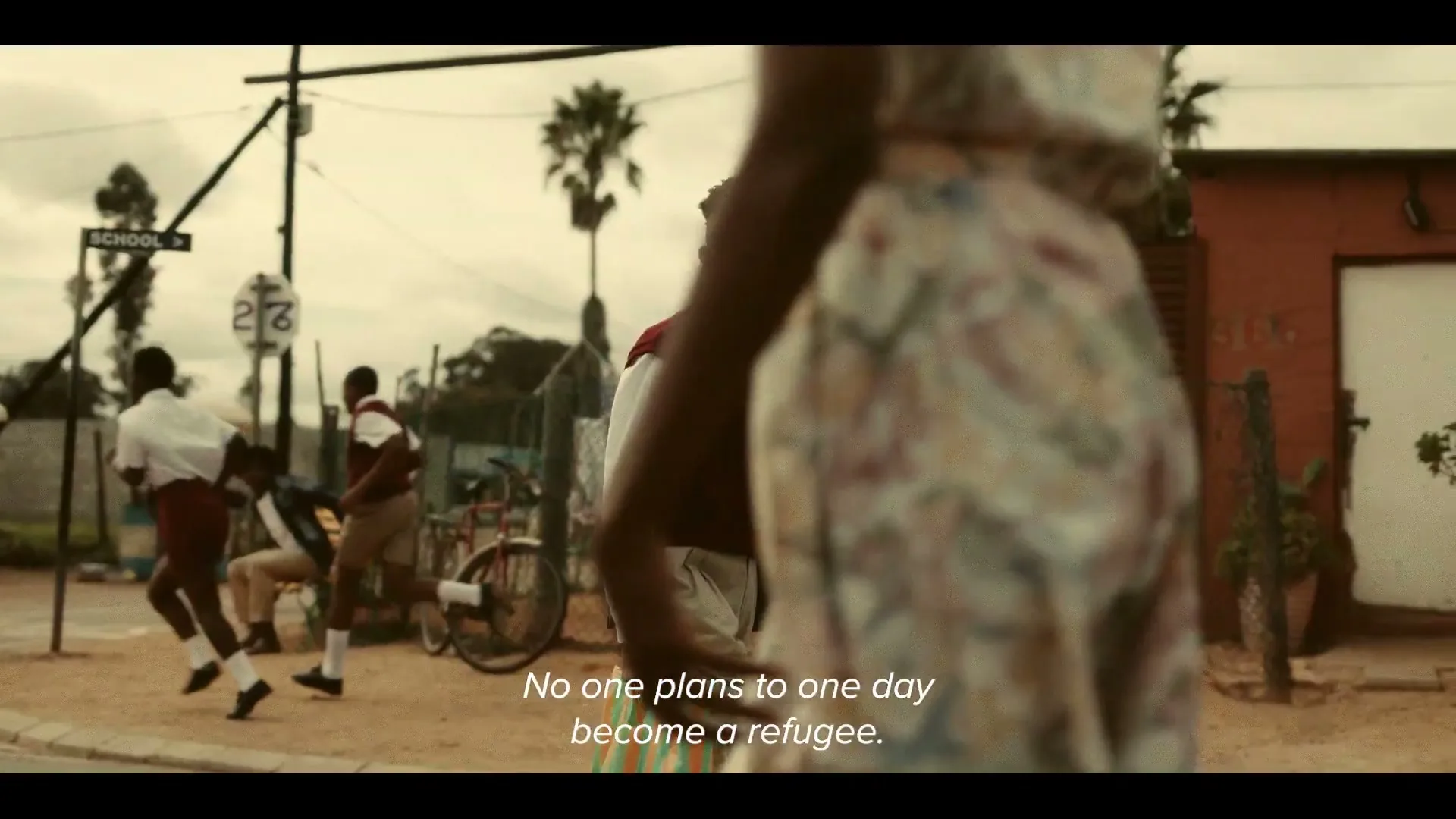 A young male student in a school uniform looks back at the camera as other students walk and play in a dusty street in a developing country.